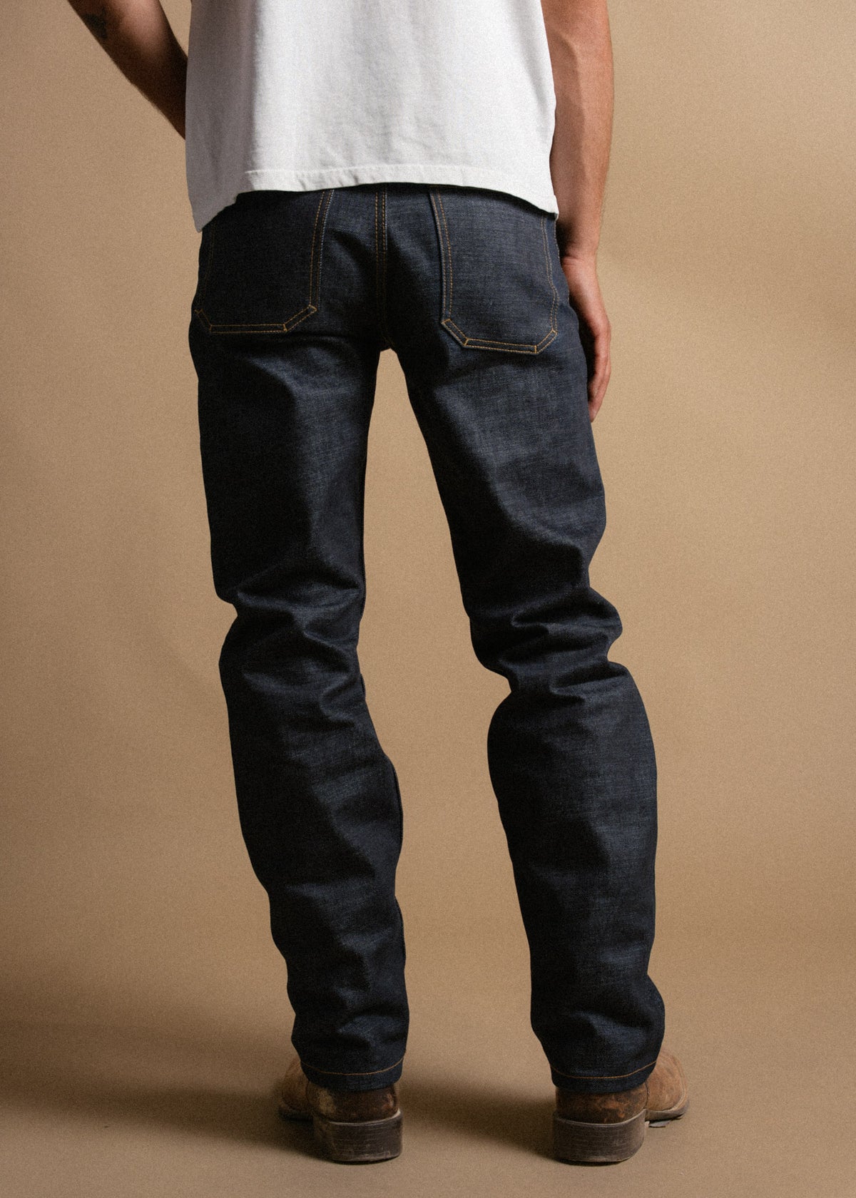 Man in a white shirt and dark jeans stands facing away on a beige background, showcasing denim fit and style.