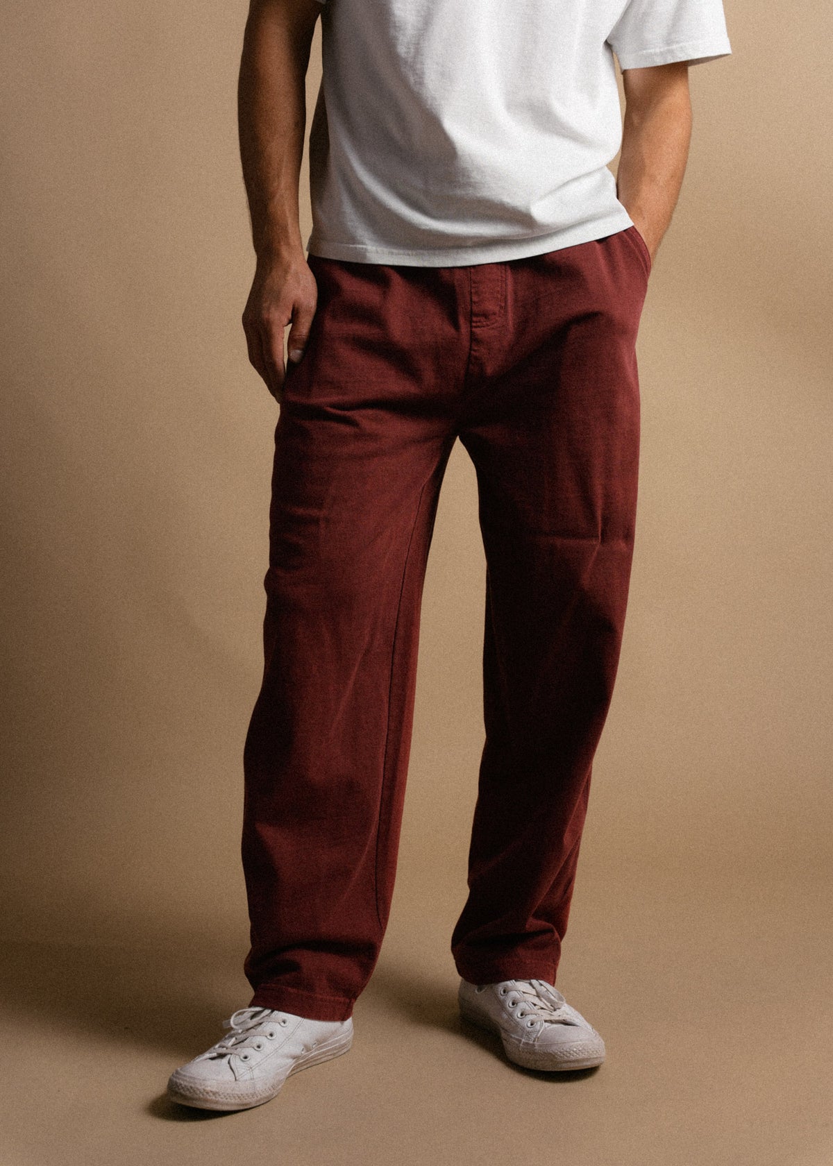 Man in white shirt and red pants stands against a beige background, hands in pockets, wearing white sneakers.