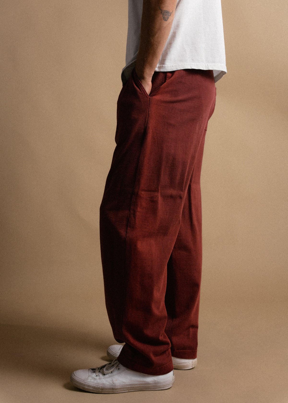 Man in red pants and white sneakers stands against a beige backdrop, hands in pockets, wearing a white shirt.