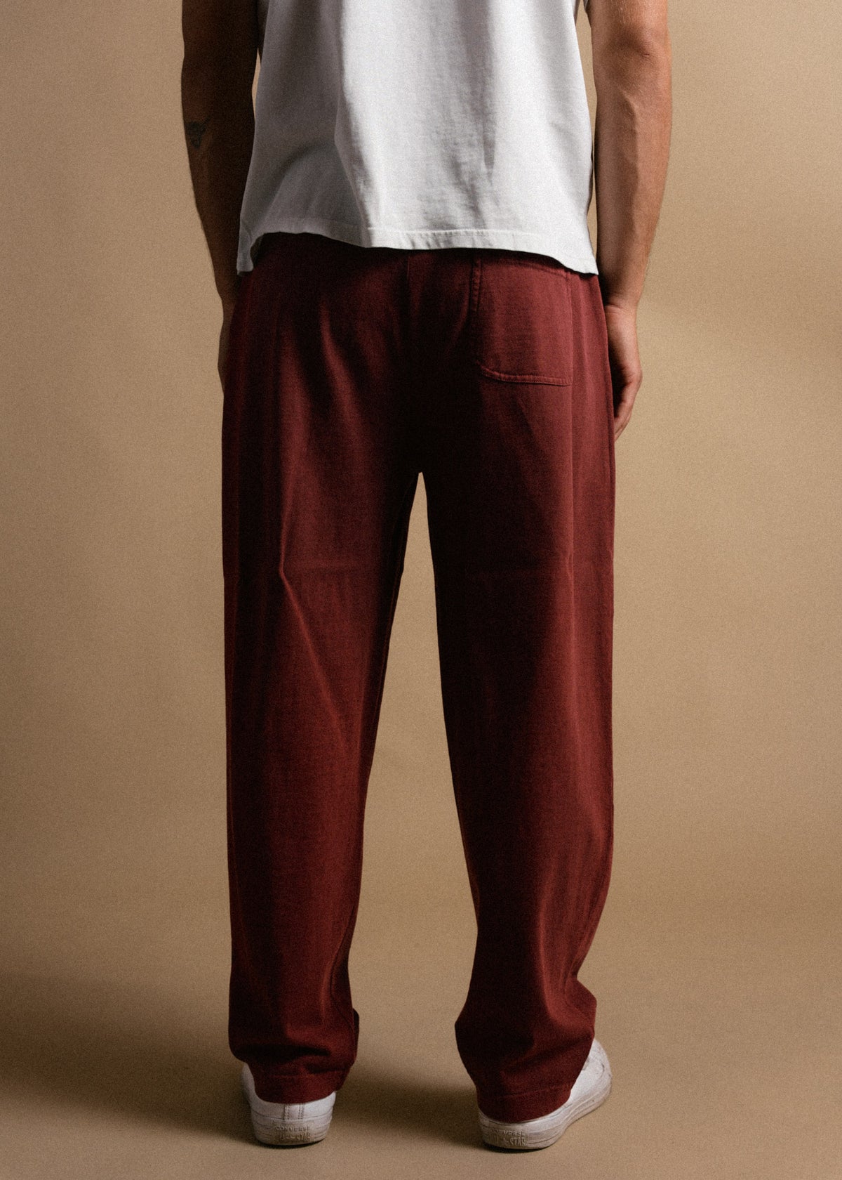 Man in white shirt and red pants stands against a beige background, facing away. Casual style with focus on clothing fit.