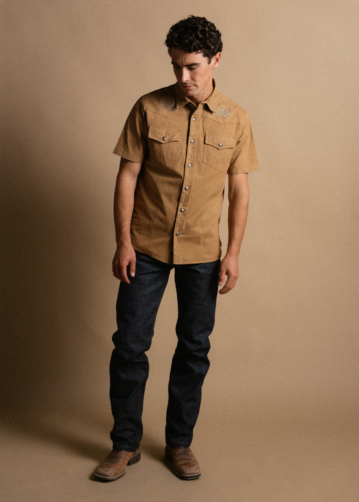 Man in a tan short-sleeve shirt with embroidery, dark jeans, and brown boots, stands against a plain beige background.