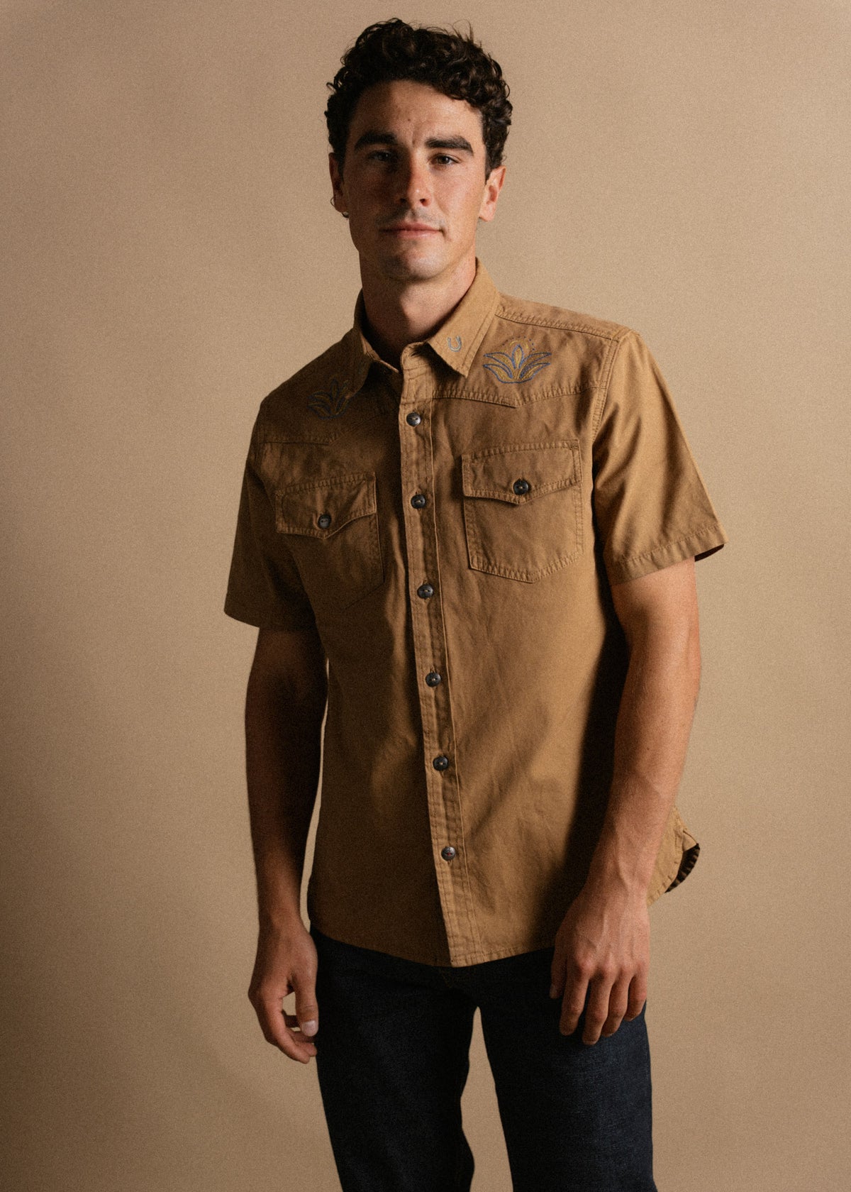 Man in a brown short-sleeve shirt with embroidered leaves stands against a beige background, looking confidently at the camera.