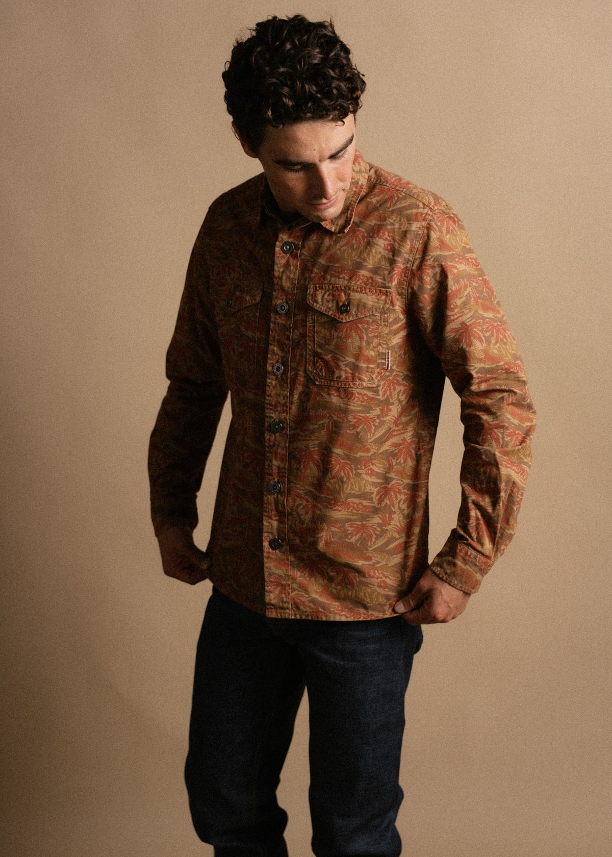 Man in a patterned brown shirt and jeans stands against a beige background, looking down with hands adjusting his shirt.