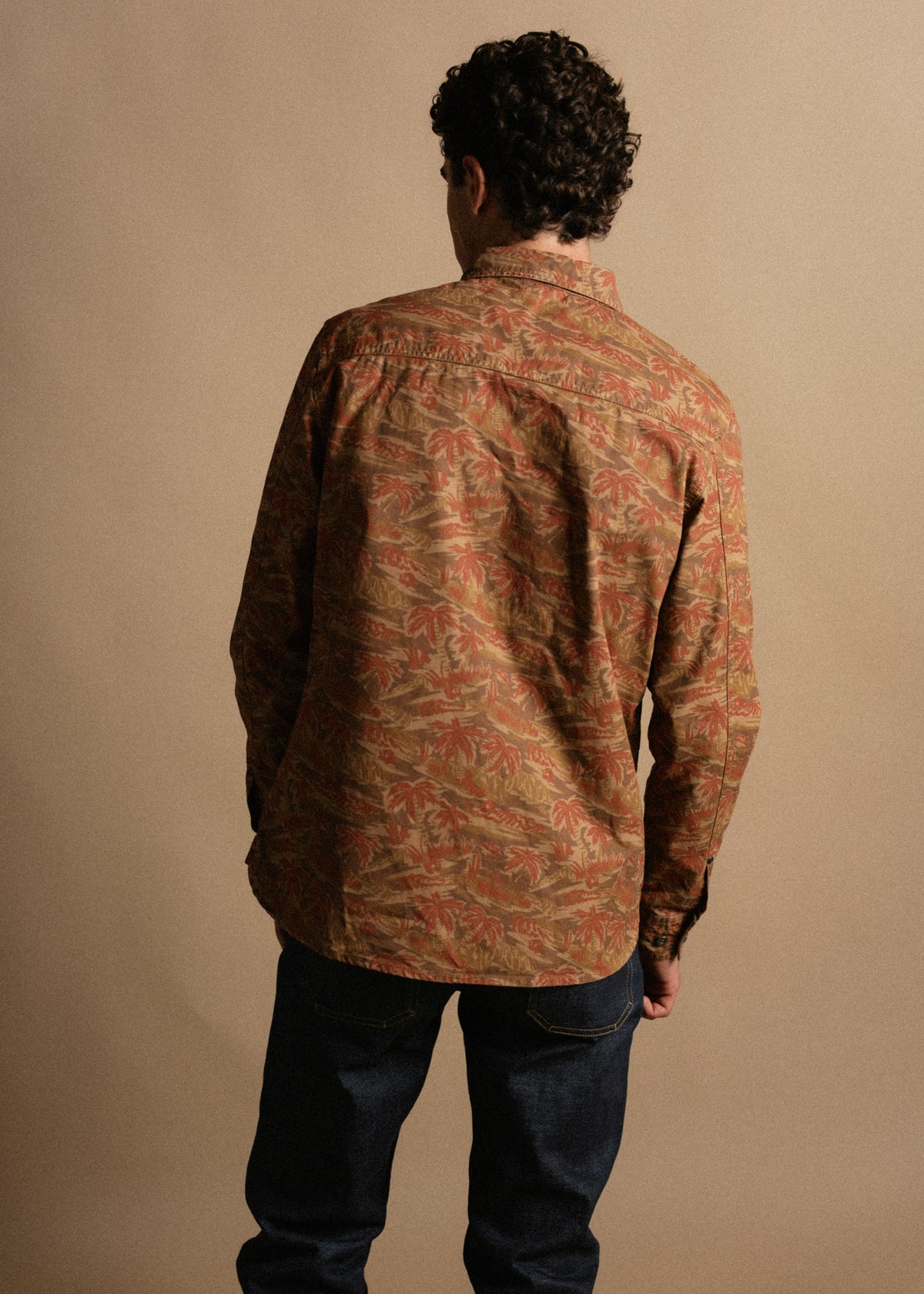 Man in a patterned shirt and jeans stands with his back to the camera against a plain background, highlighting the shirt's design.