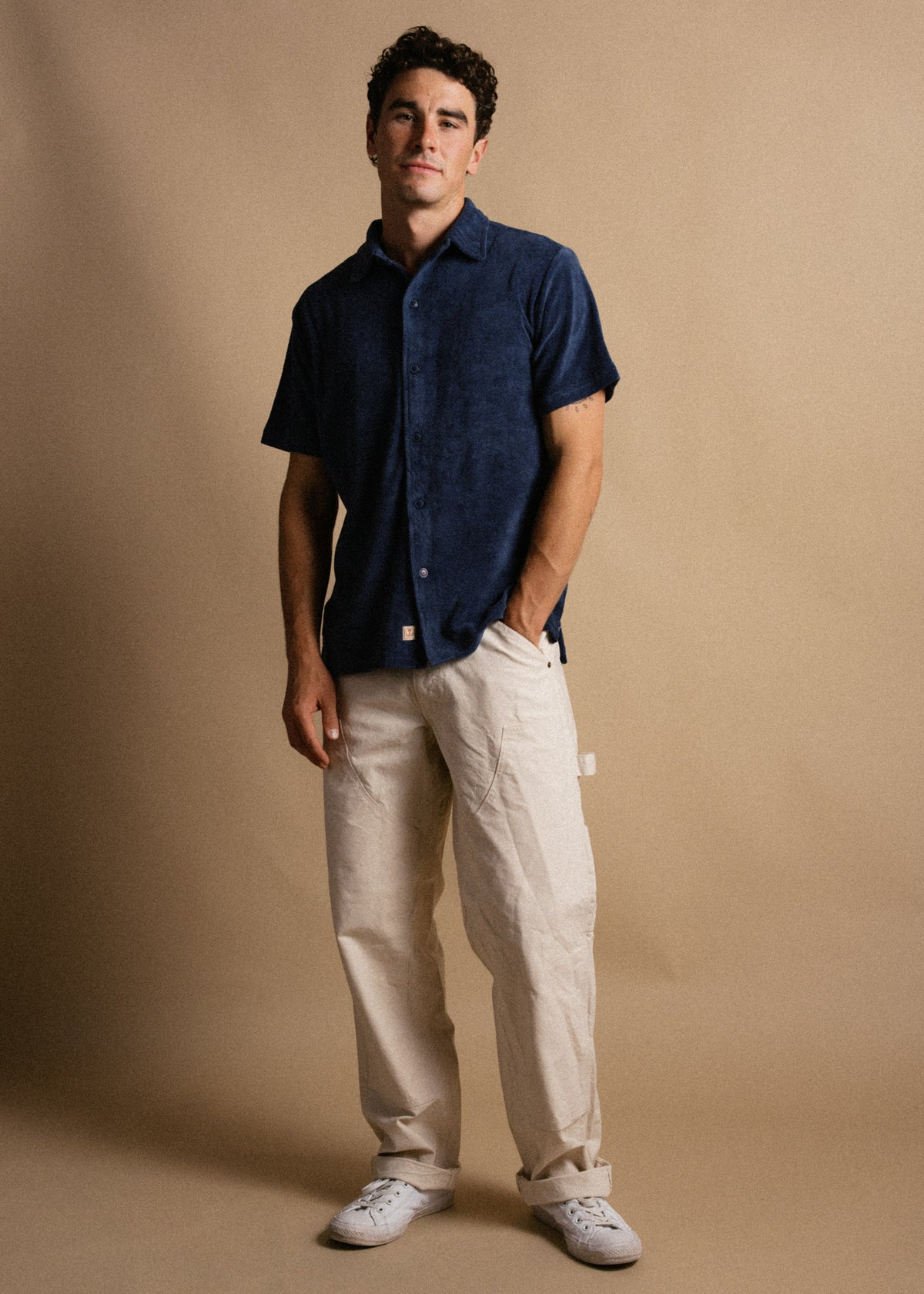 Man in a blue shirt and beige pants stands against a tan background, exuding casual style with a relaxed posture and white sneakers.