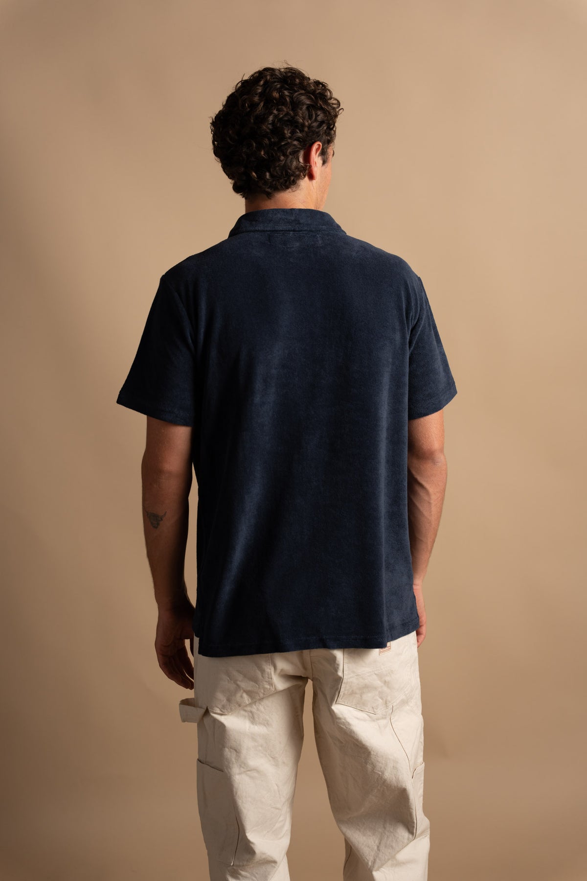 Man in navy shirt and beige pants stands with back to camera against a neutral background, showcasing casual fashion style.