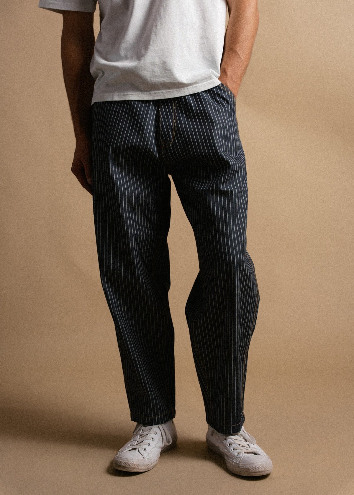 Man in a white shirt and pinstriped pants stands casually with hands in pockets, wearing white sneakers against a beige backdrop.