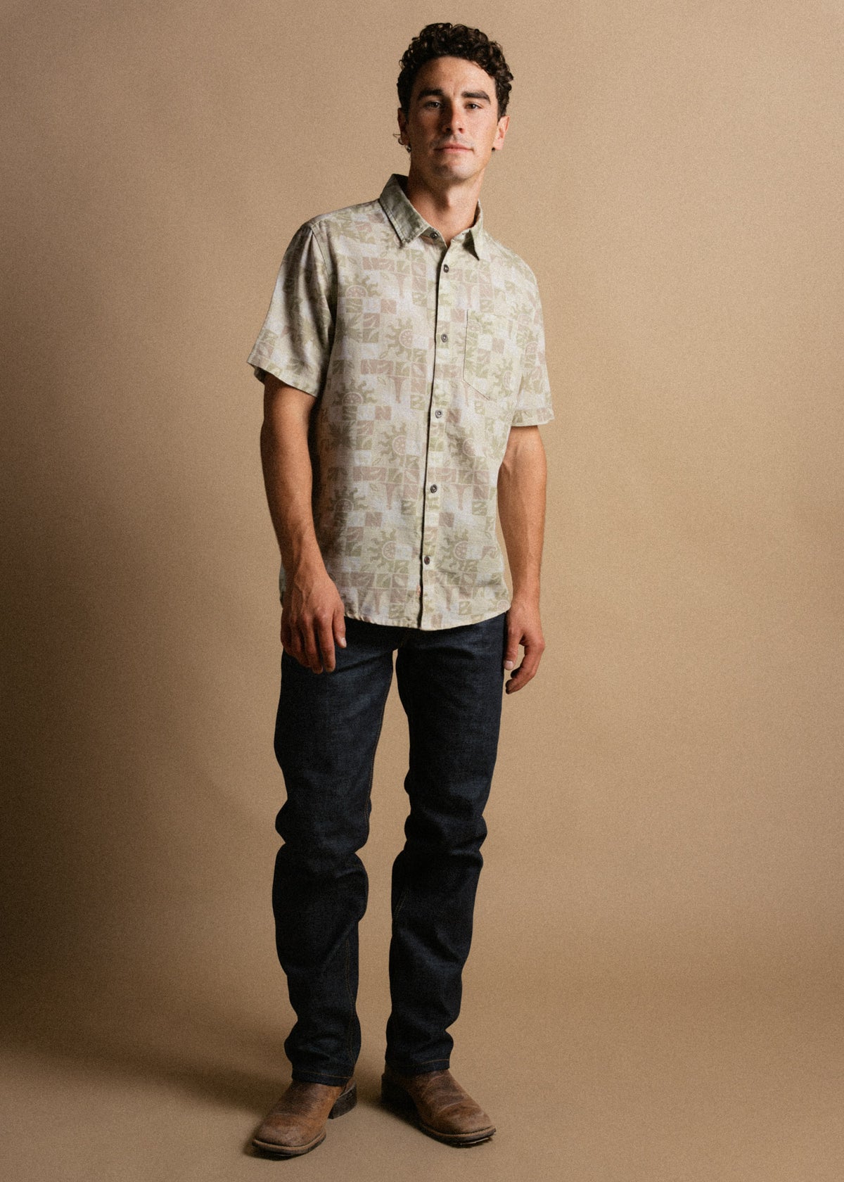 Man in a patterned short-sleeve shirt, dark jeans, and brown boots stands against a beige background, looking confidently ahead.