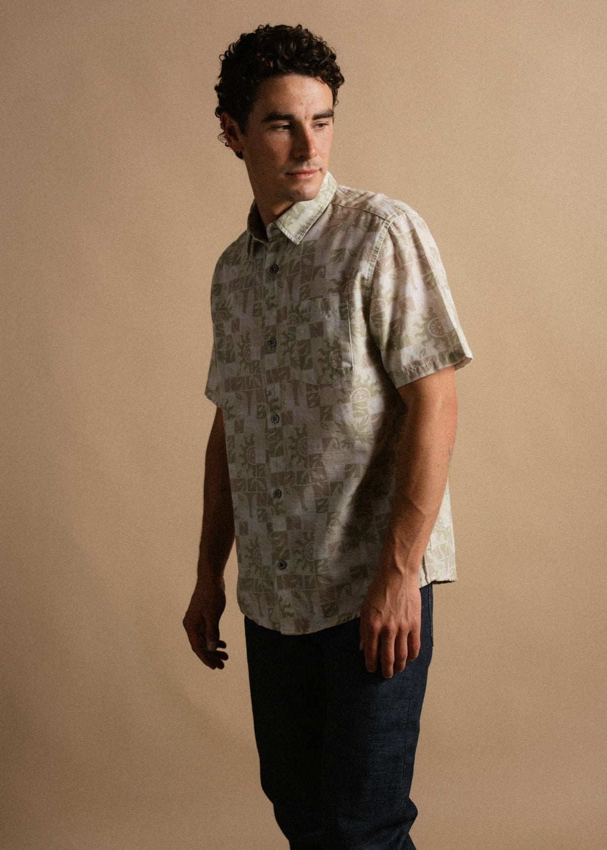 Man in patterned short-sleeve shirt and jeans stands against a beige background, looking to the side with a thoughtful expression.