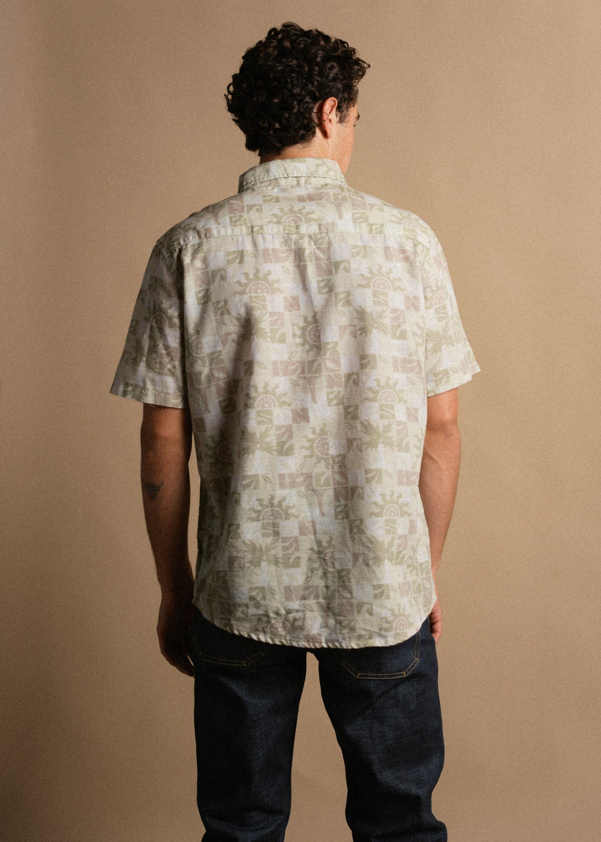 Man in patterned short-sleeve shirt and jeans, standing with back to camera against a beige background, showcasing casual style.