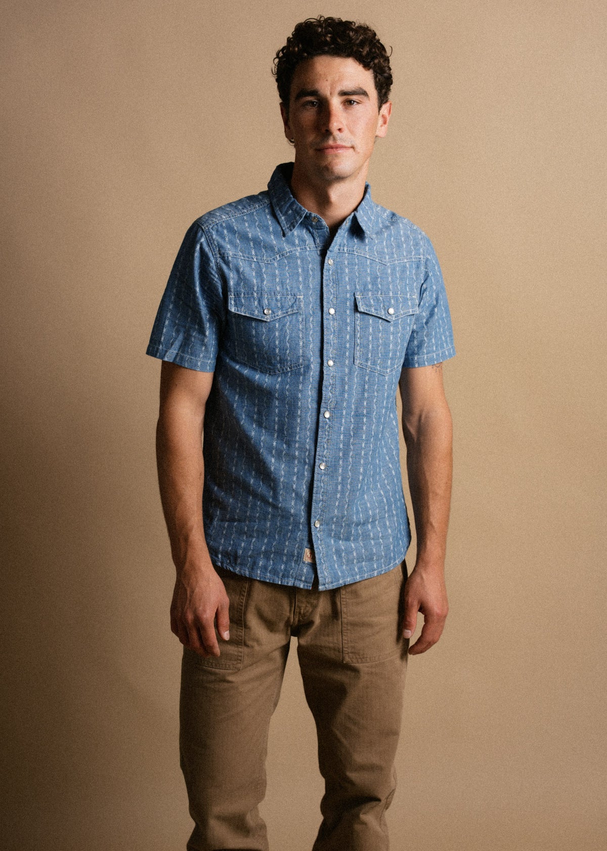 Man in a blue short-sleeve button-up shirt and brown pants stands against a beige background, looking directly at the camera.