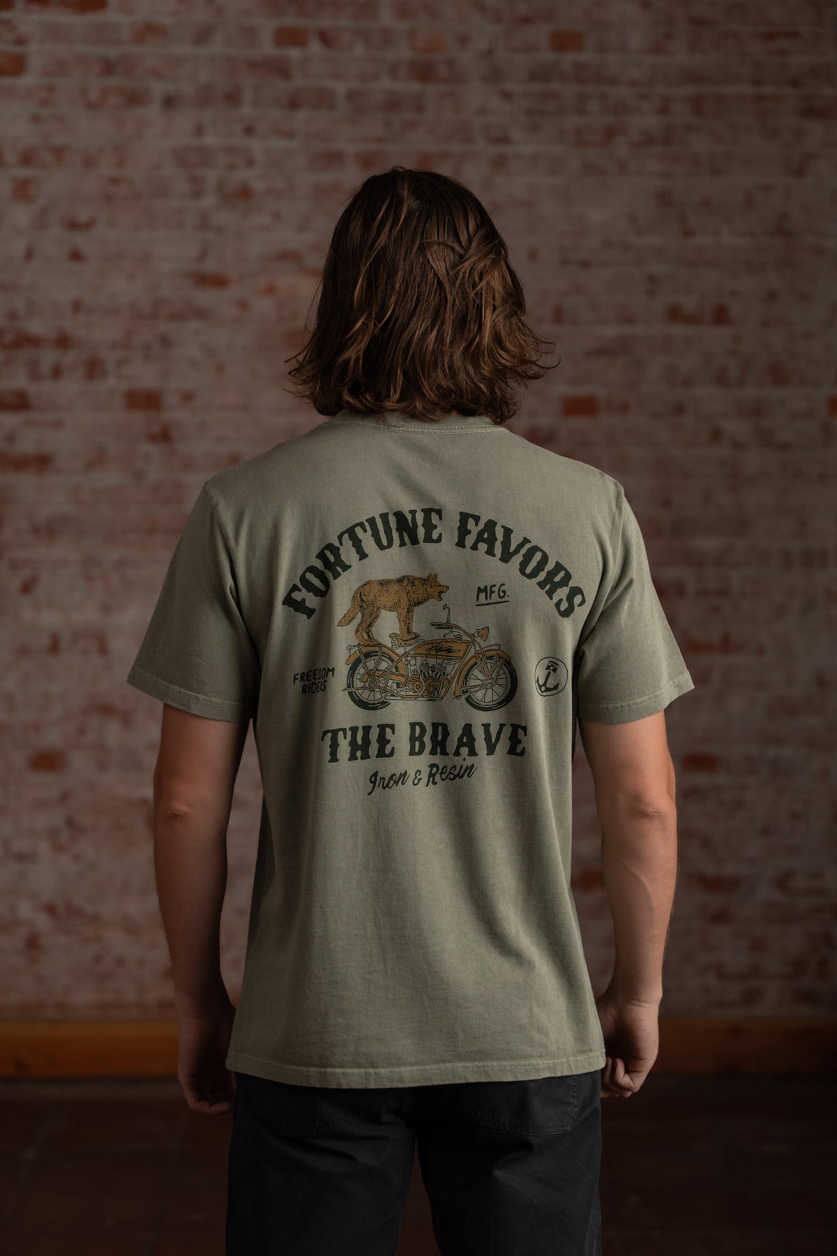Man in olive shirt with "Fortune Favors the Brave" text and motorcycle graphic, standing against a brick wall.