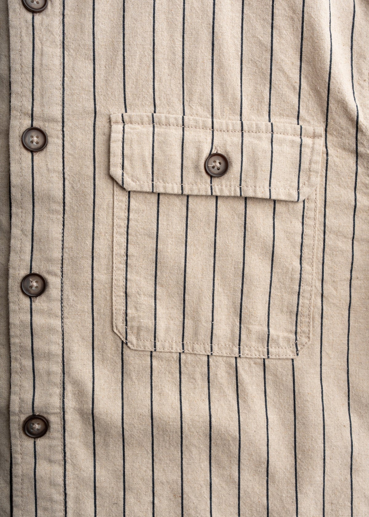 Close-up of a beige shirt with thin black stripes, featuring brown buttons and a pocket with a buttoned flap.