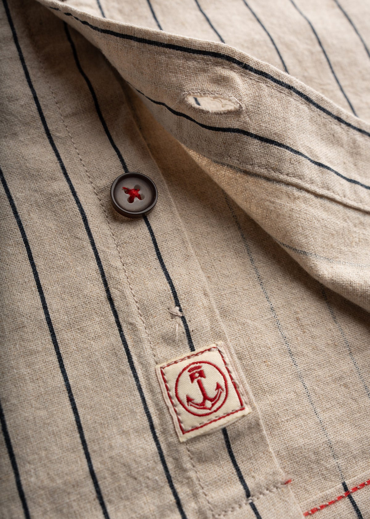 Close-up of a beige striped shirt with a red anchor logo patch and a button detail, highlighting nautical style.
