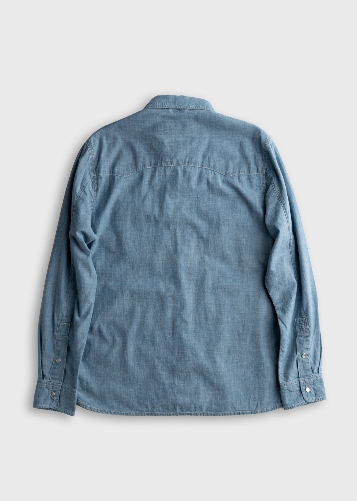 A light blue denim shirt is laid flat, showcasing its back with long sleeves and buttoned cuffs, on a plain gray background.