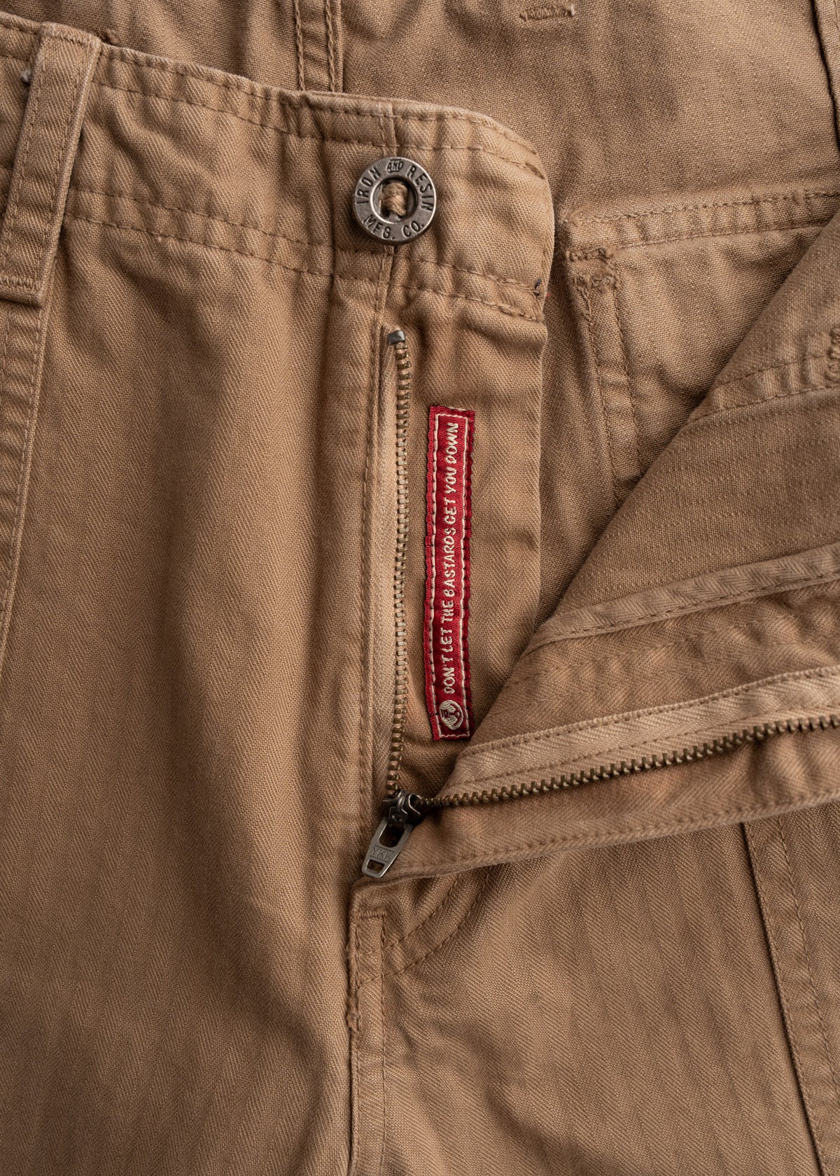 Close-up of tan pants with a metal button and zipper. Red label reads "DON'T LET THE BASTARDS GET YOU DOWN."