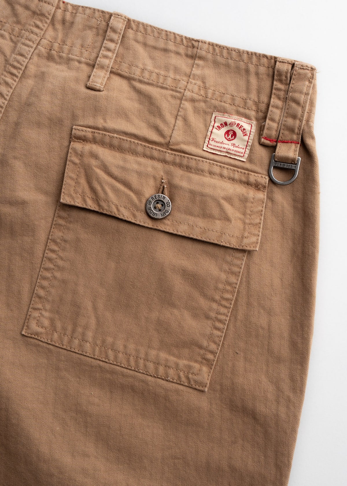 Close-up of brown pants with a buttoned pocket, Iron and Resin label, and metal D-ring detail, showcasing rugged style.