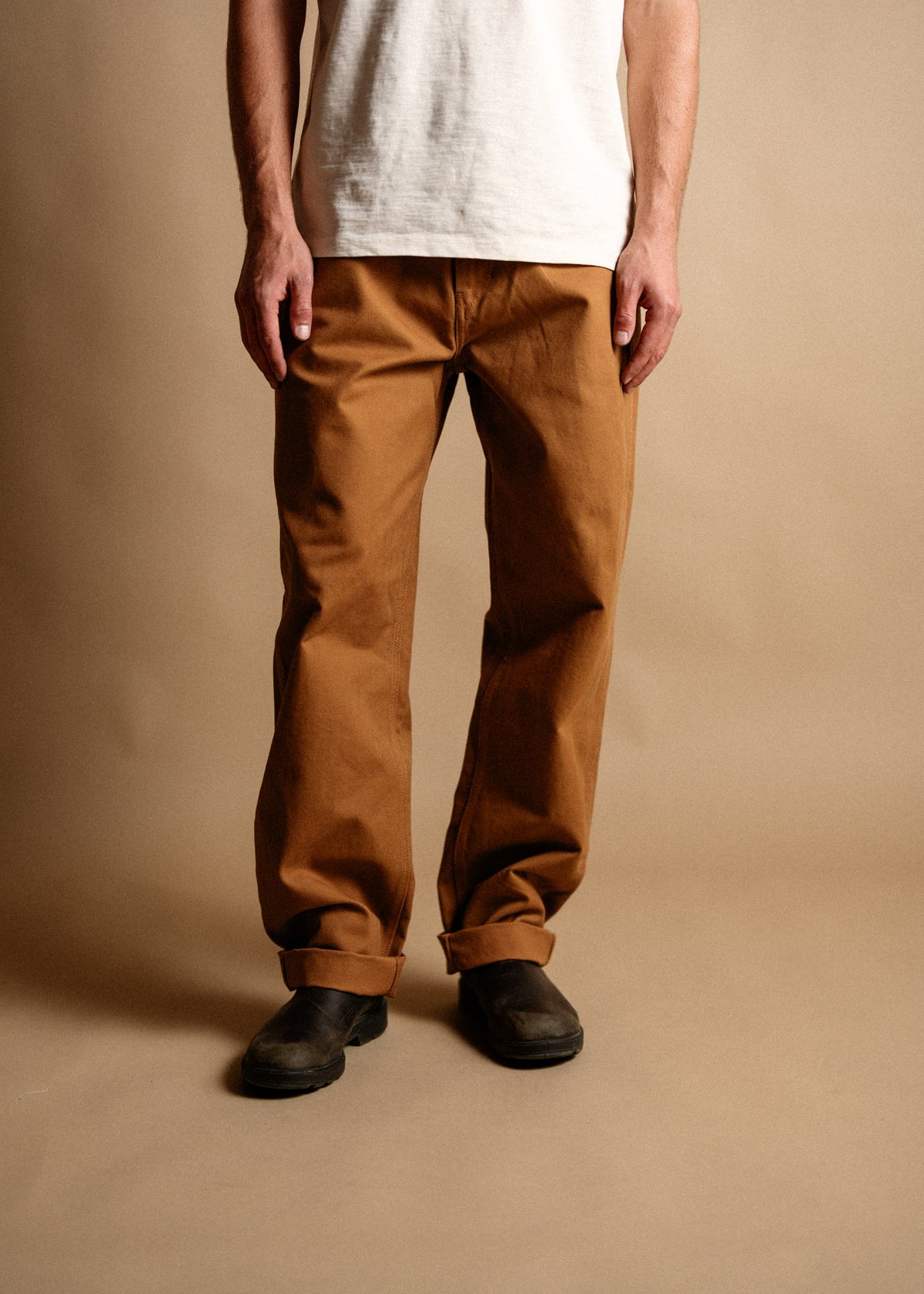 Man in white shirt, brown pants, and black shoes stands against a beige background, showcasing casual fashion style.