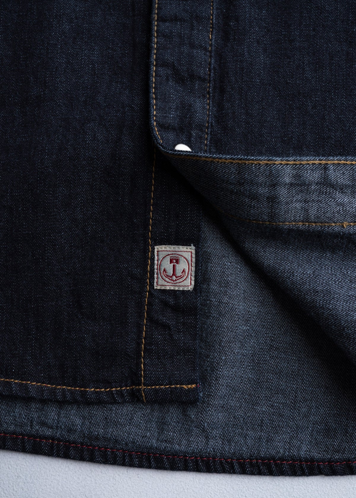 Close-up of dark denim fabric with visible stitching and a small red anchor logo tag, partially folded to reveal inner texture.