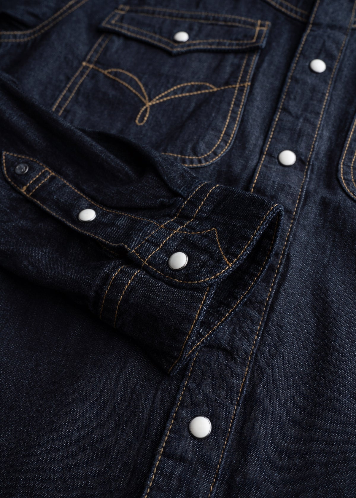 Close-up of a dark denim shirt with white snap buttons and contrast stitching, highlighting its detailed pocket and cuff design.