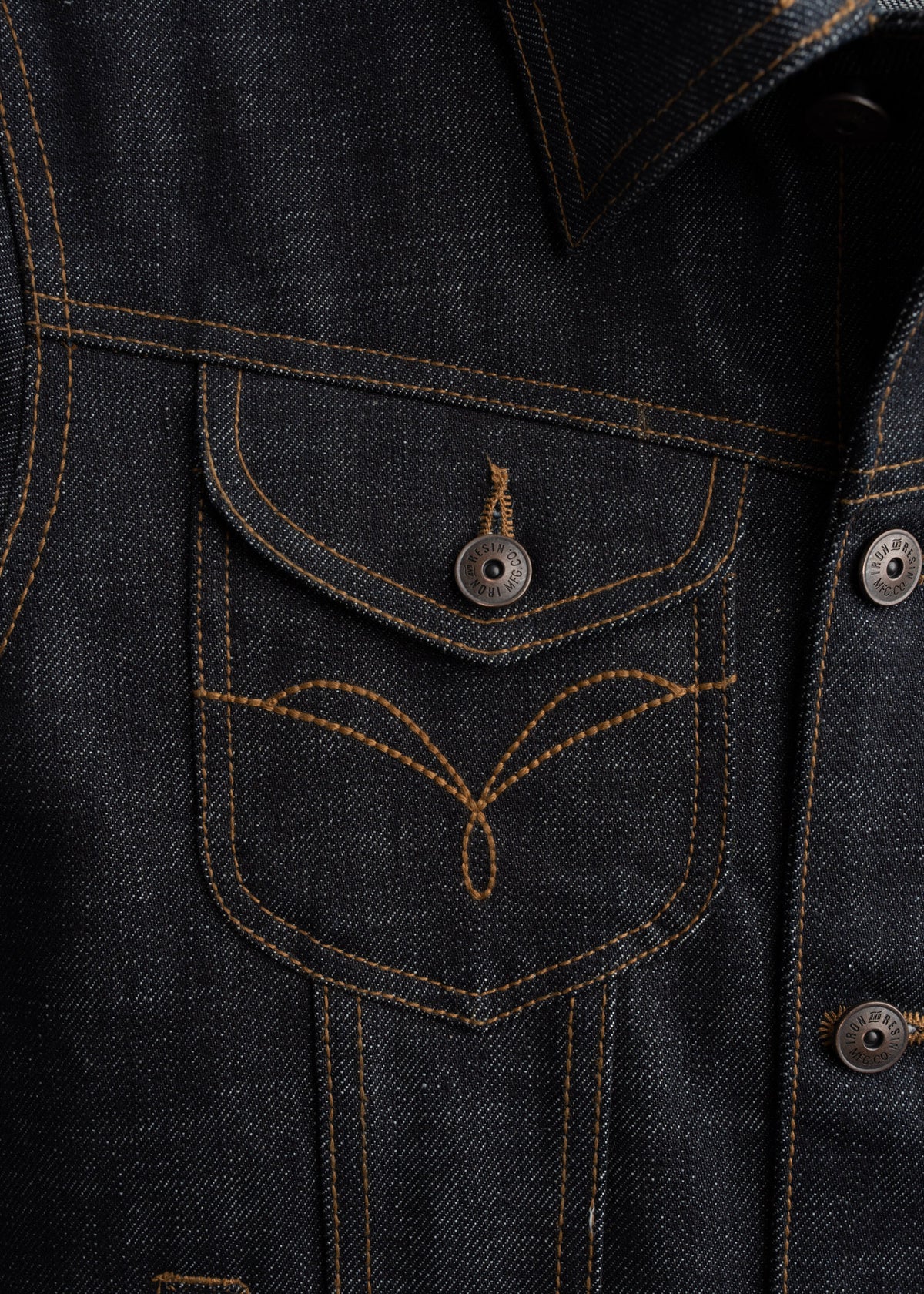 Close-up of a denim jacket pocket with orange stitching and metal buttons, showcasing detailed craftsmanship and texture.