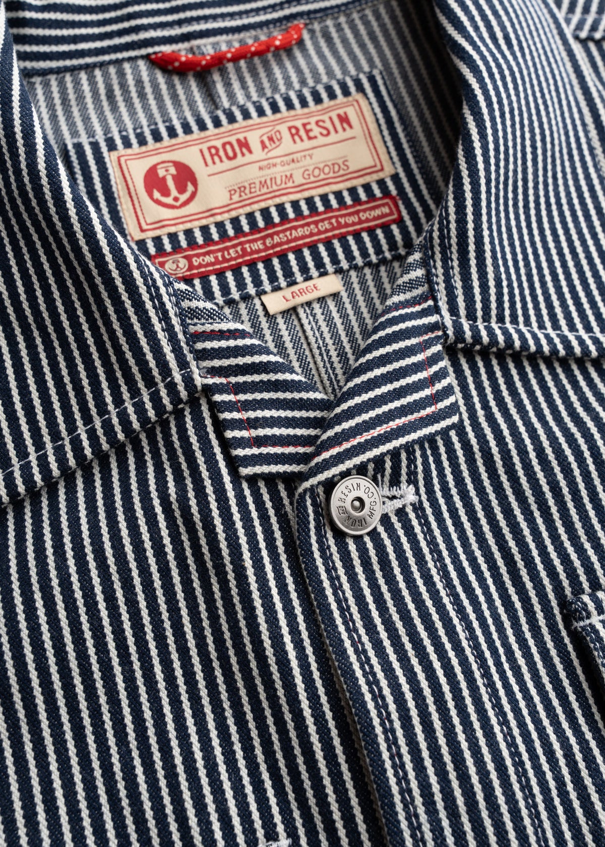 Close-up of a striped shirt with "Iron and Resin" label, featuring a button and red stitching detail. Size tag reads "Large."