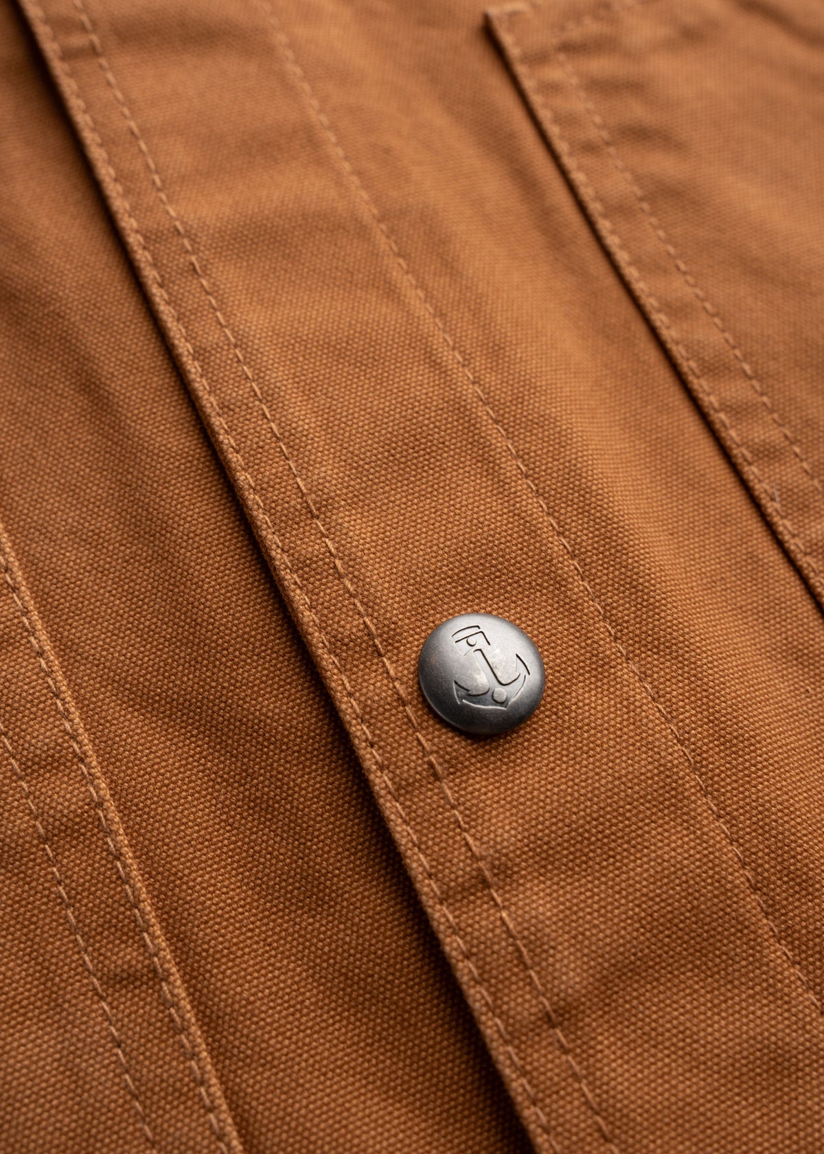 Close-up of a brown fabric with a metal button featuring an anchor design, highlighting texture and stitching detail.