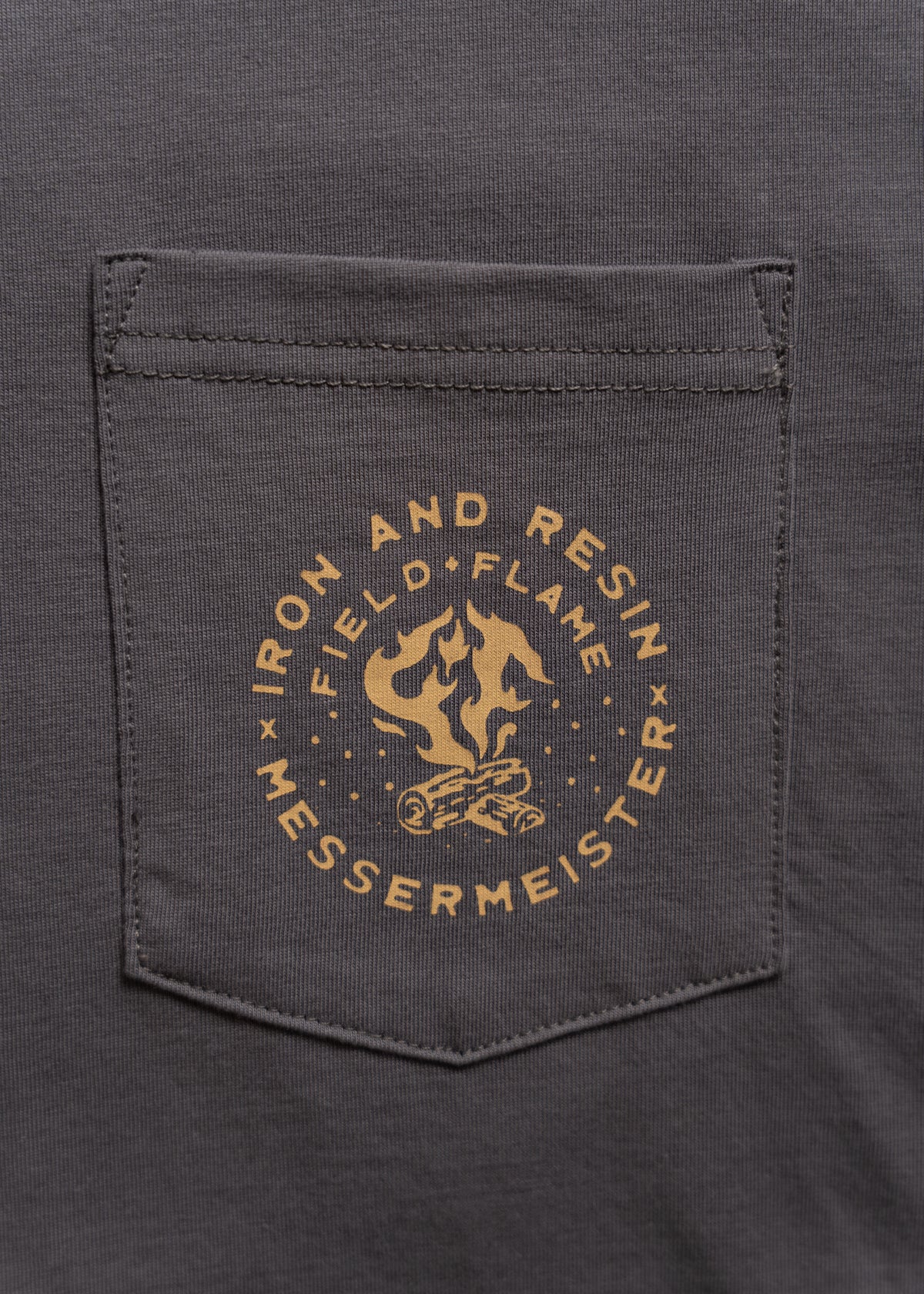 Gray shirt pocket with "Iron and Resin Field Flame Messermeister" logo in yellow, featuring a flame and log design.