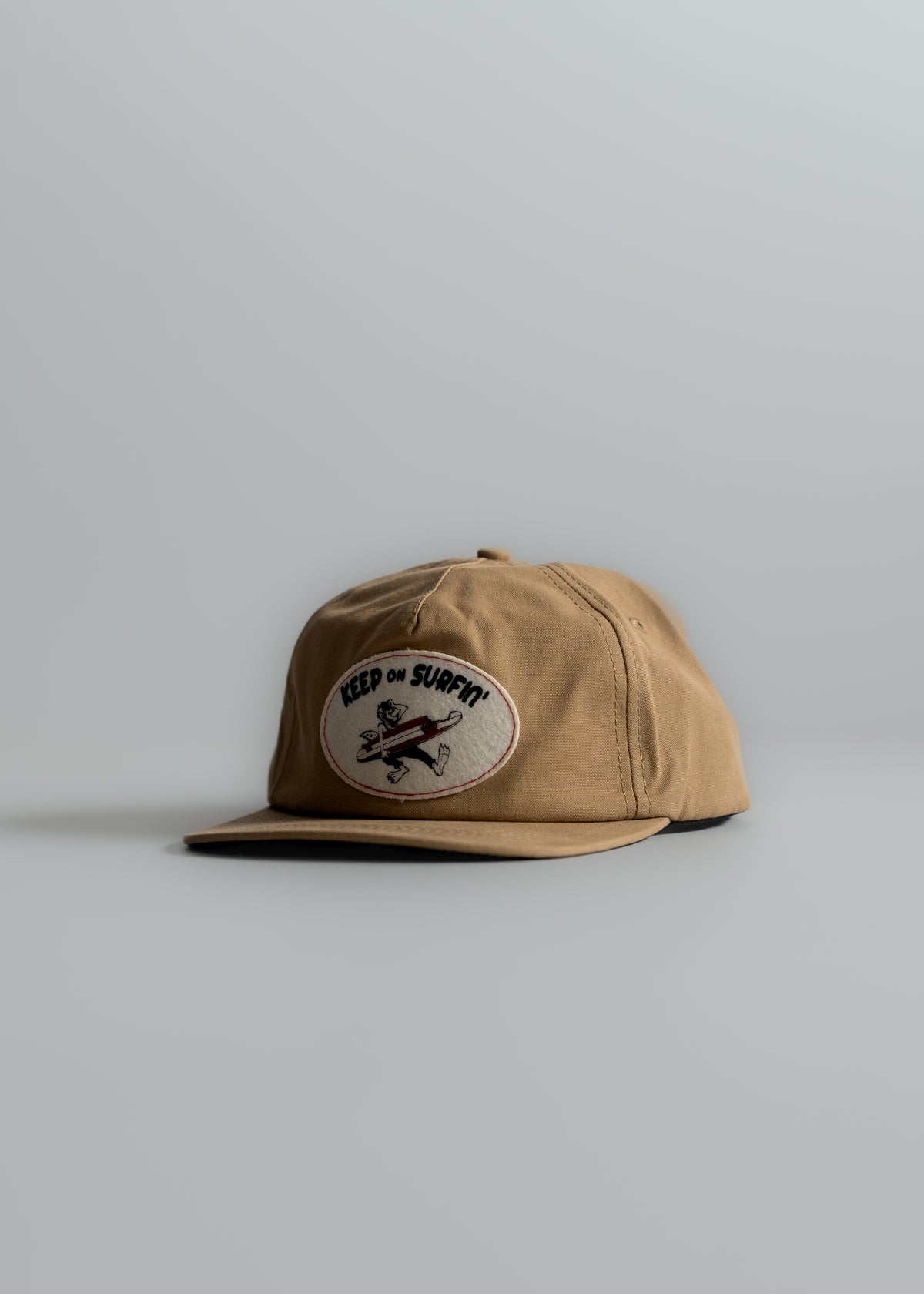 Tan cap with "Keep on Surfin'" patch, featuring a cartoon surfer, on a neutral background. Simple and stylish design.