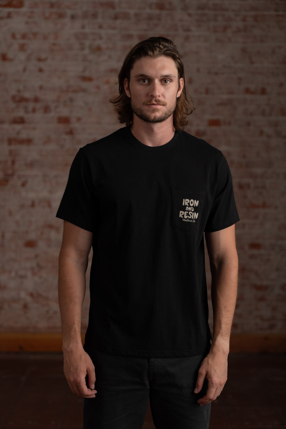 Man in black "Iron and Resin" shirt stands against a brick wall, looking forward. Casual and relaxed pose.
