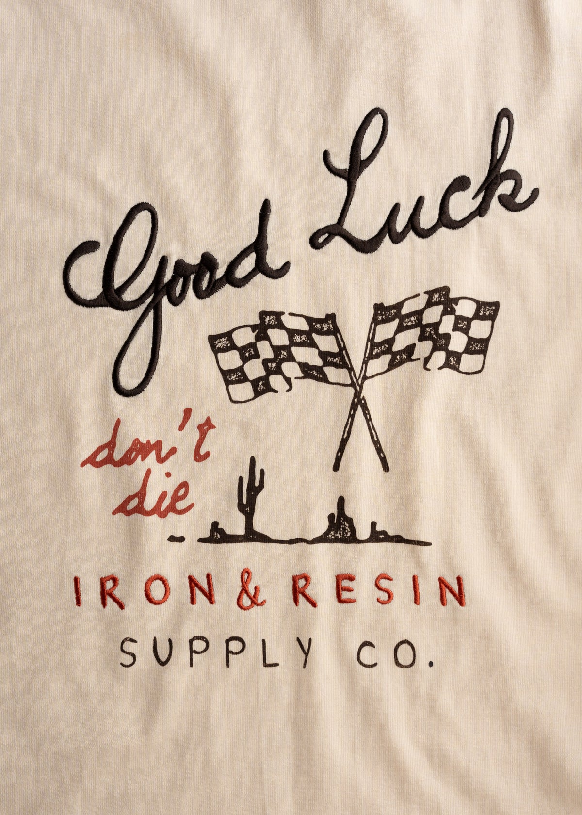 "Good Luck" and "don't die" text with checkered flags, cactus, and "IRON & RESIN SUPPLY CO." logo on a beige fabric background.