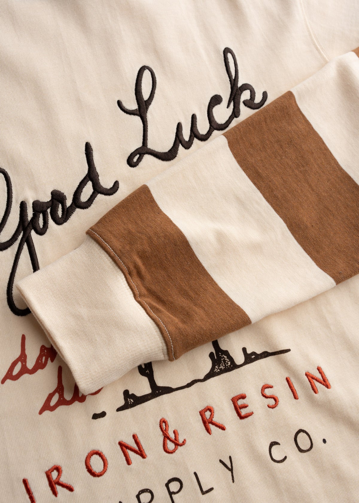 Close-up of a beige shirt with "Good Luck" text and Iron & Resin Supply Co. logo, featuring brown and white striped sleeve.