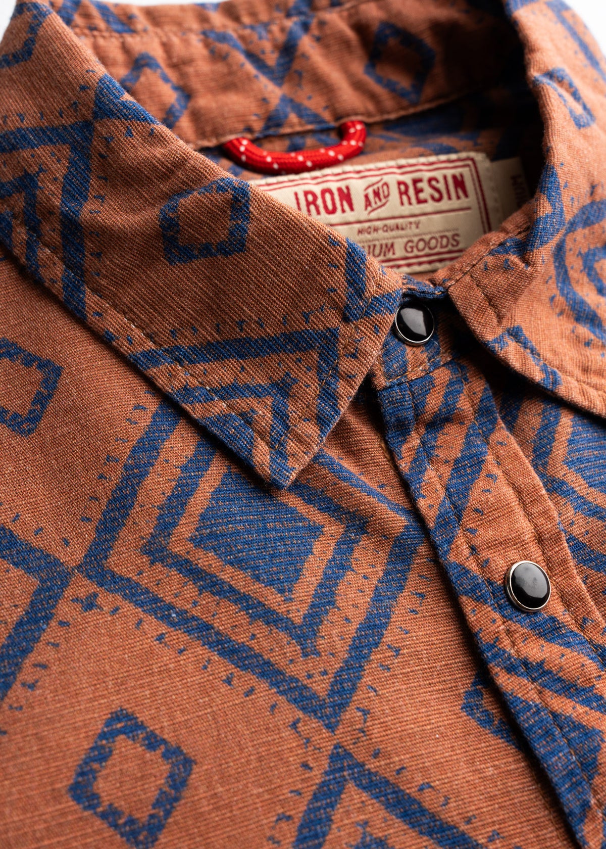 Close-up of a brown shirt with blue geometric patterns and black buttons, featuring an Iron and Resin label inside the collar.