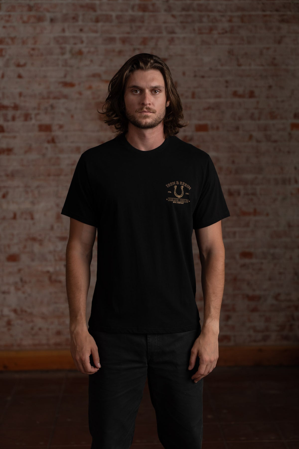 Man in a black t-shirt stands against a brick wall, looking forward. The shirt has a small logo on the left chest.