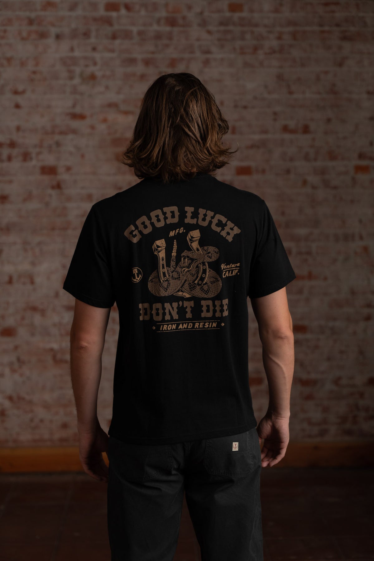 Man with long hair wearing a black "Good Luck Don't Die" T-shirt stands against a brick wall, facing away from the camera.