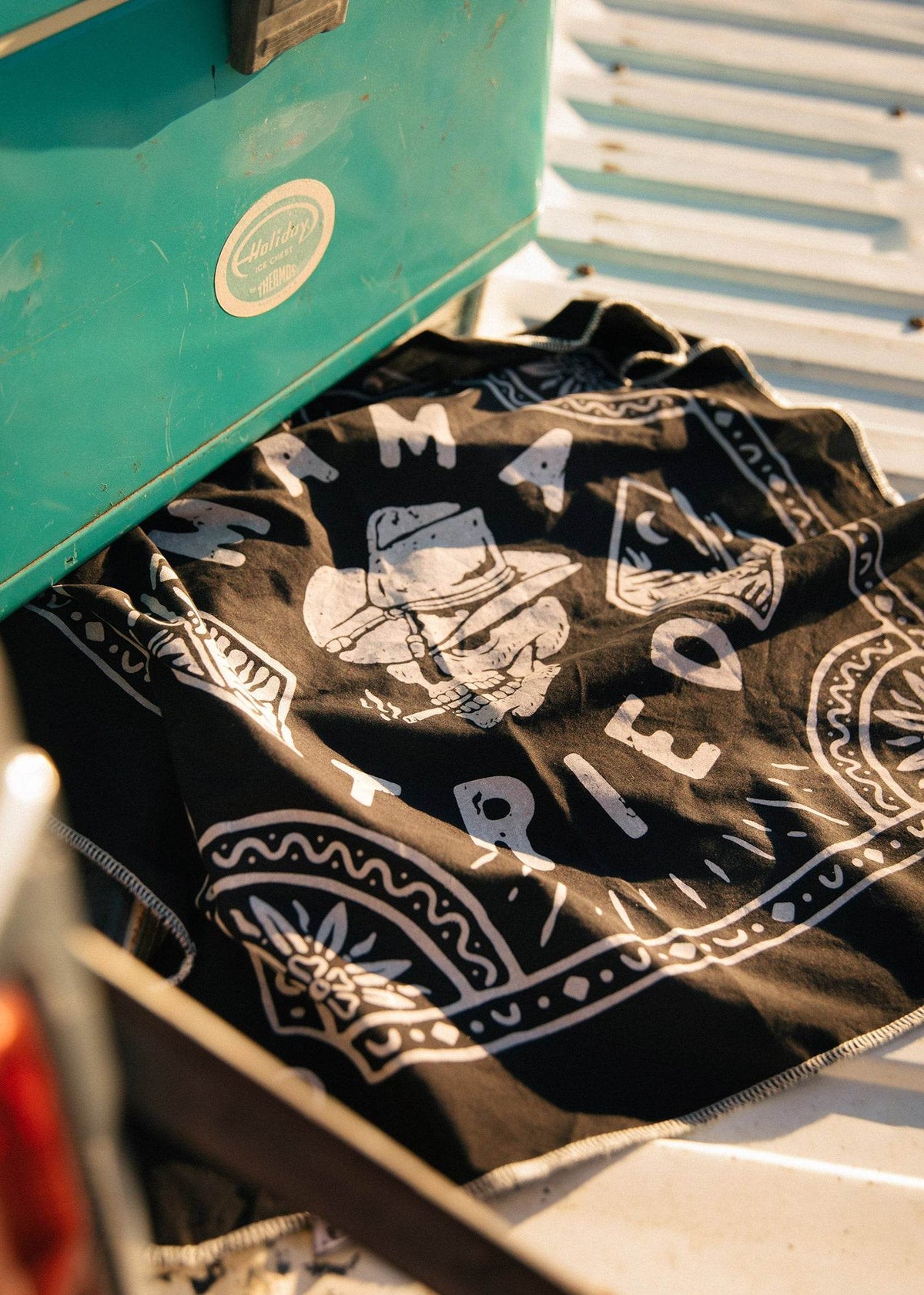A teal cooler sits on a truck bed beside a black bandana with "Mama Tried" and a cowboy design in white.