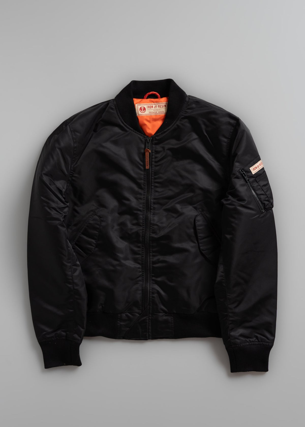 Black bomber jacket with orange lining and zip pocket on sleeve, displayed on a gray background.