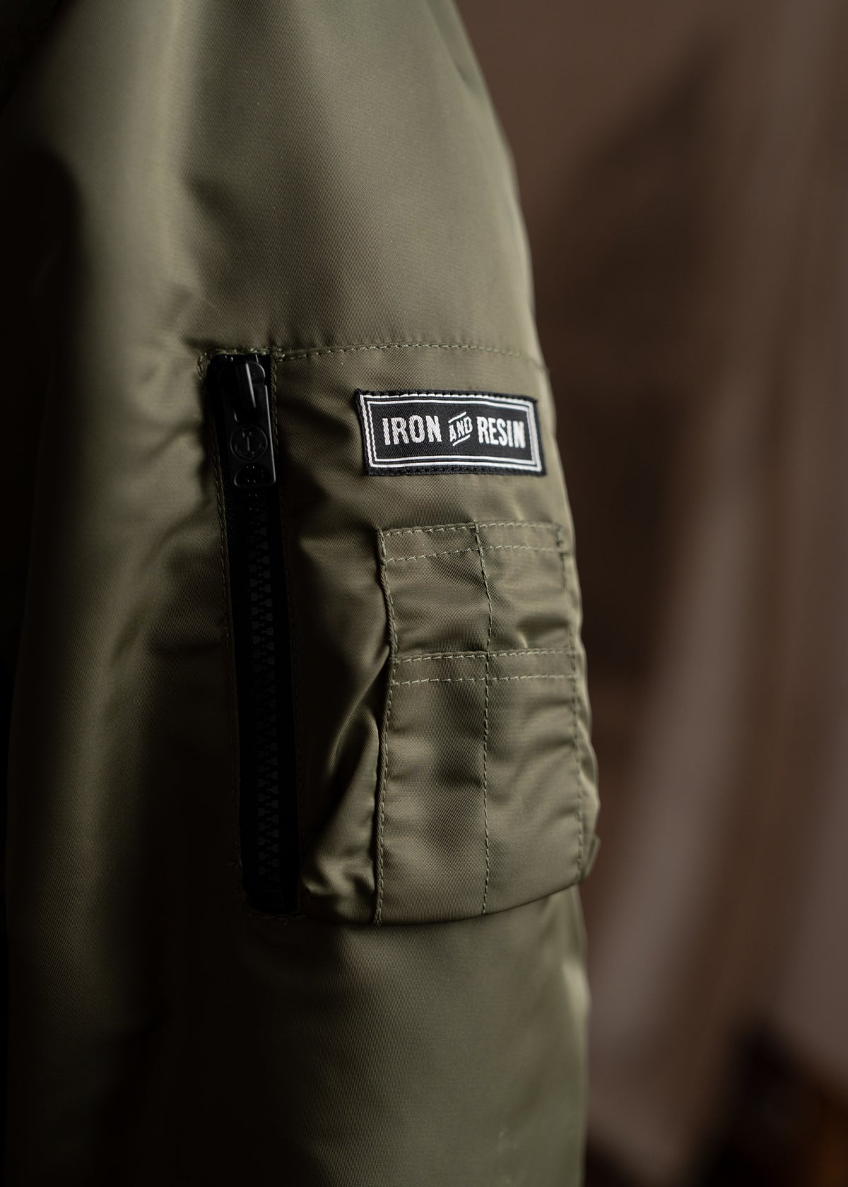 Close-up of an olive green jacket sleeve with a black zipper and "Iron and Resin" patch, highlighting its rugged style.