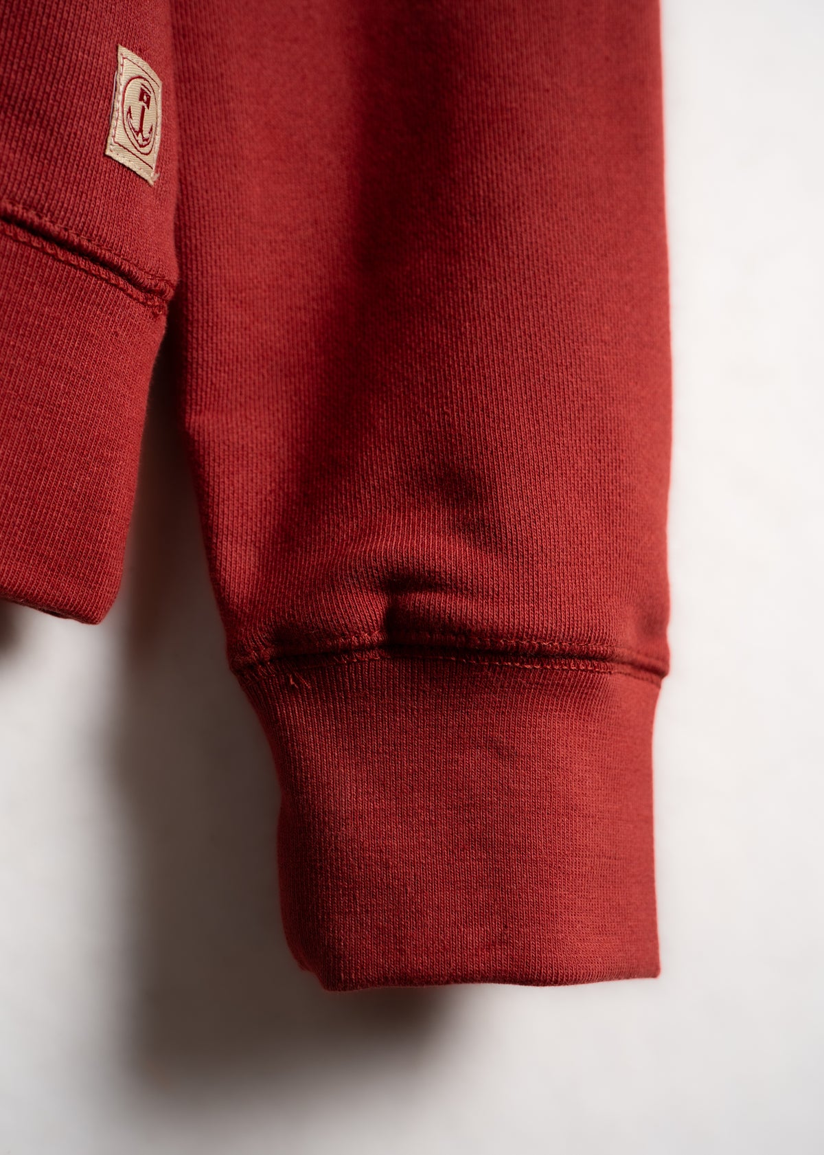 Close-up of a red sweatshirt sleeve with a small anchor patch on a white background, highlighting the texture and stitching.