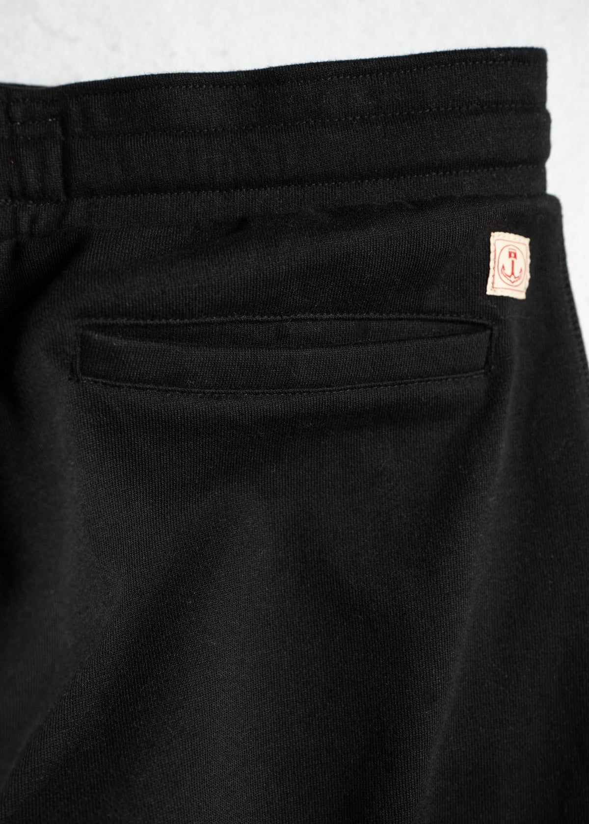 Close-up of black pants with a back pocket and a small anchor logo tag on the waistband, highlighting the design details.