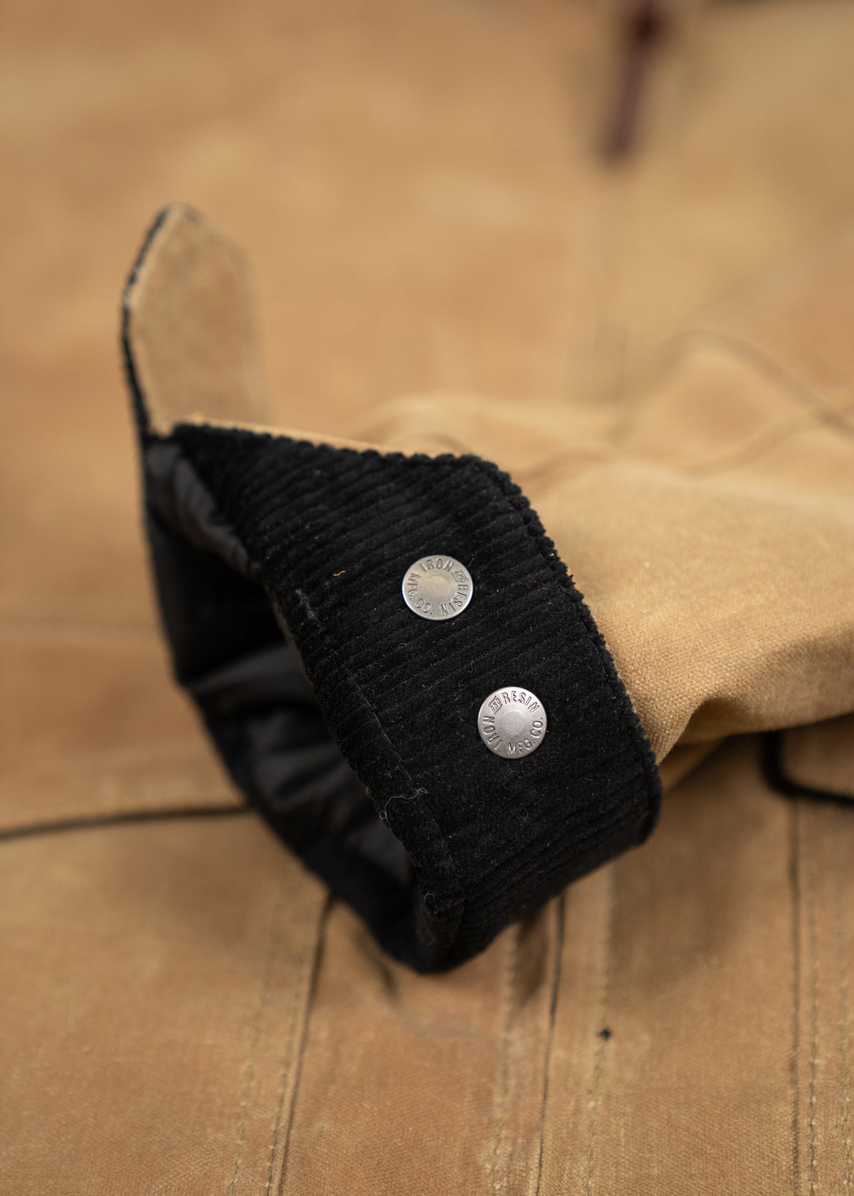 Close-up of a tan jacket sleeve with black corduroy cuff and two silver buttons, showcasing detailed stitching and texture.