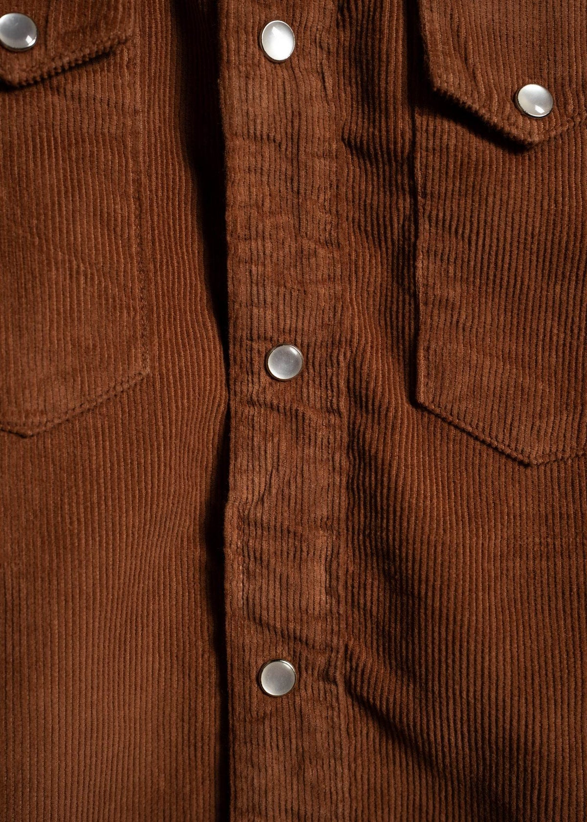 Close-up of a brown corduroy shirt with silver snap buttons and two chest pockets, showcasing its textured fabric and design.