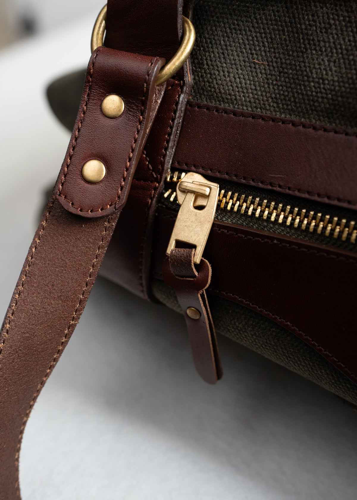 Close-up of a brown leather bag with a brass zipper and strap, showcasing detailed stitching and metal accents on a fabric background.