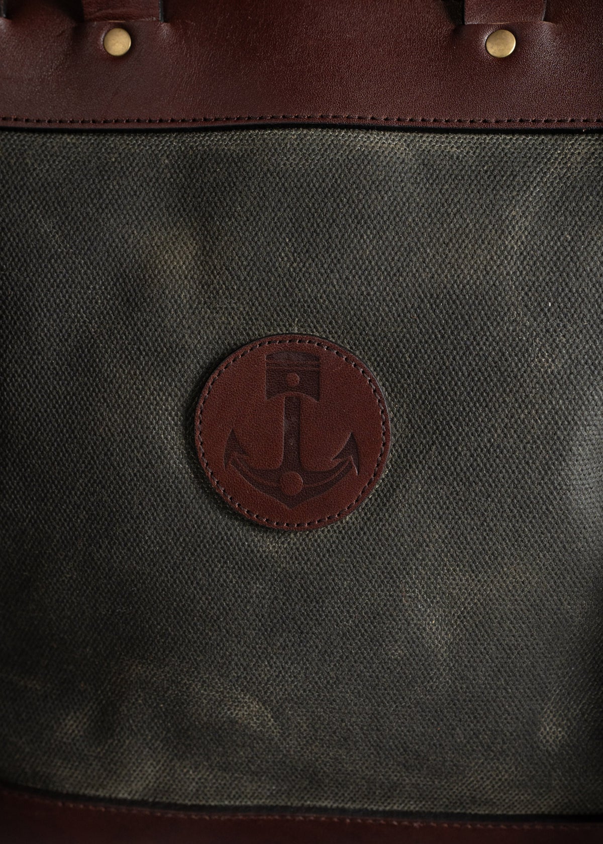 Brown leather bag with anchor emblem on canvas fabric, featuring brass rivets. Classic nautical design and sturdy craftsmanship.