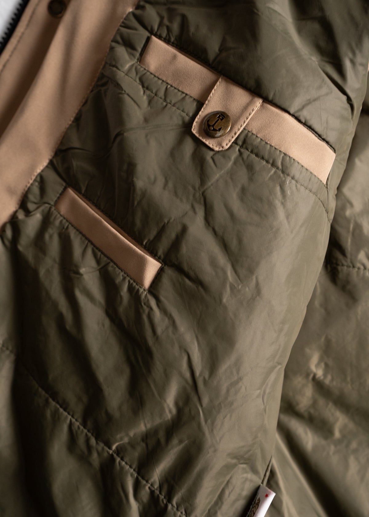 Close-up of a green jacket with beige accents, featuring a button with an anchor design and a neatly stitched pocket detail.