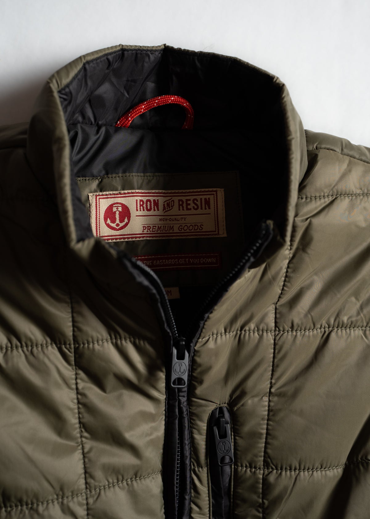 Olive quilted jacket with black lining, red hanger loop, and "Iron and Resin" label, featuring zippers with anchor logos.
