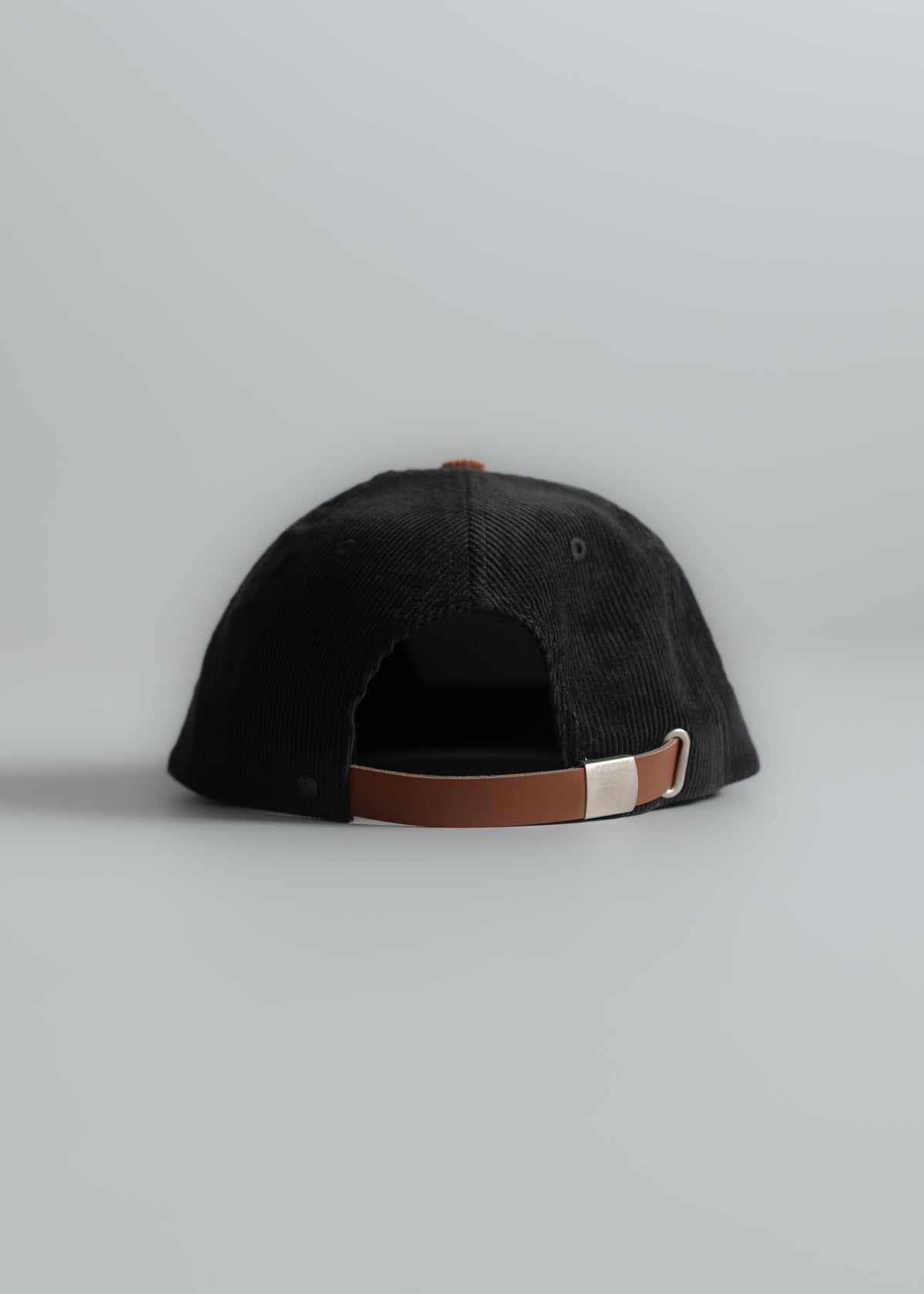 Black corduroy cap with brown leather strap and metal buckle, viewed from the back on a light gray background.