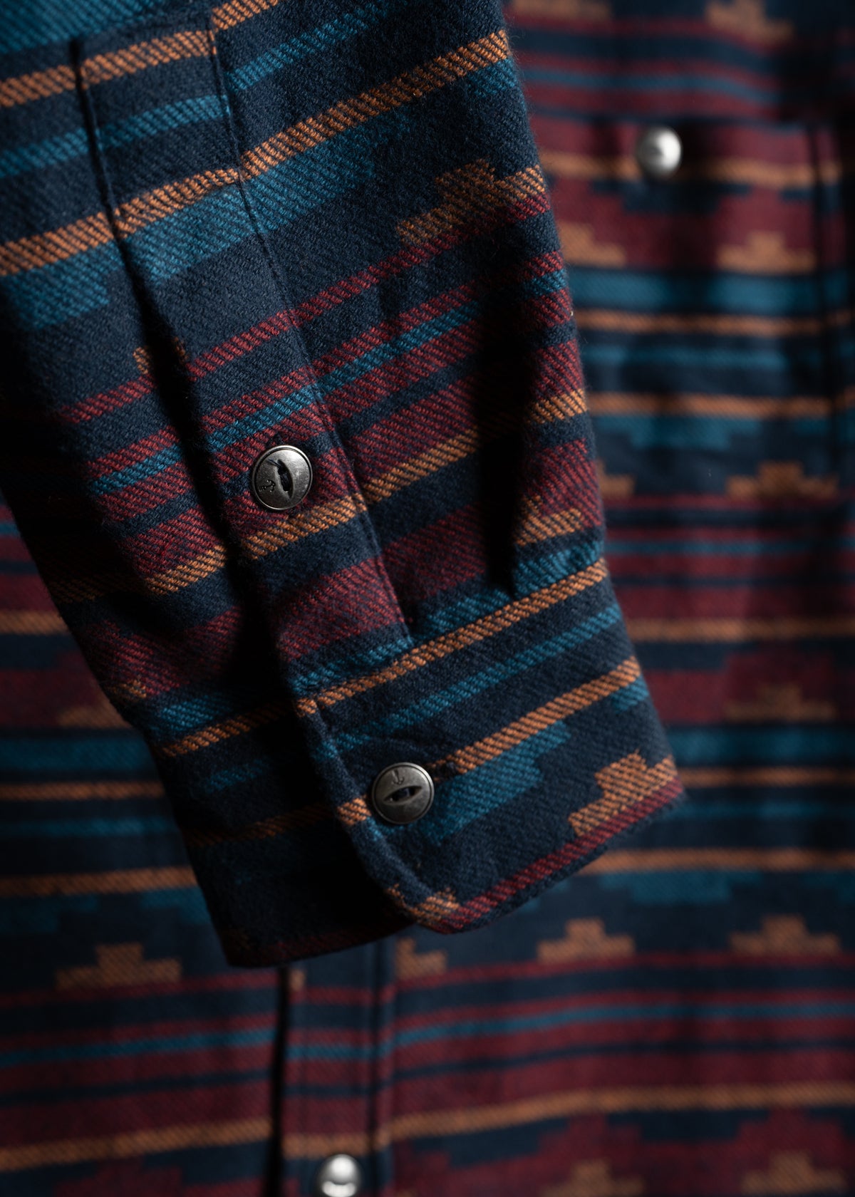 Close-up of a flannel shirt sleeve with a colorful, geometric pattern and metal buttons, highlighting its detailed texture.