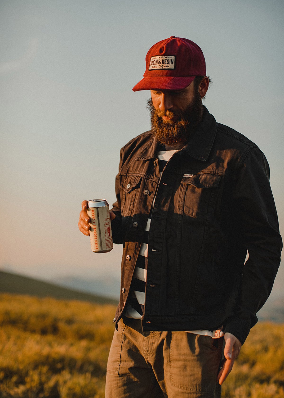 Man in a red cap and denim jacket holds a can outdoors at sunset, standing on grassy terrain with a serene expression.