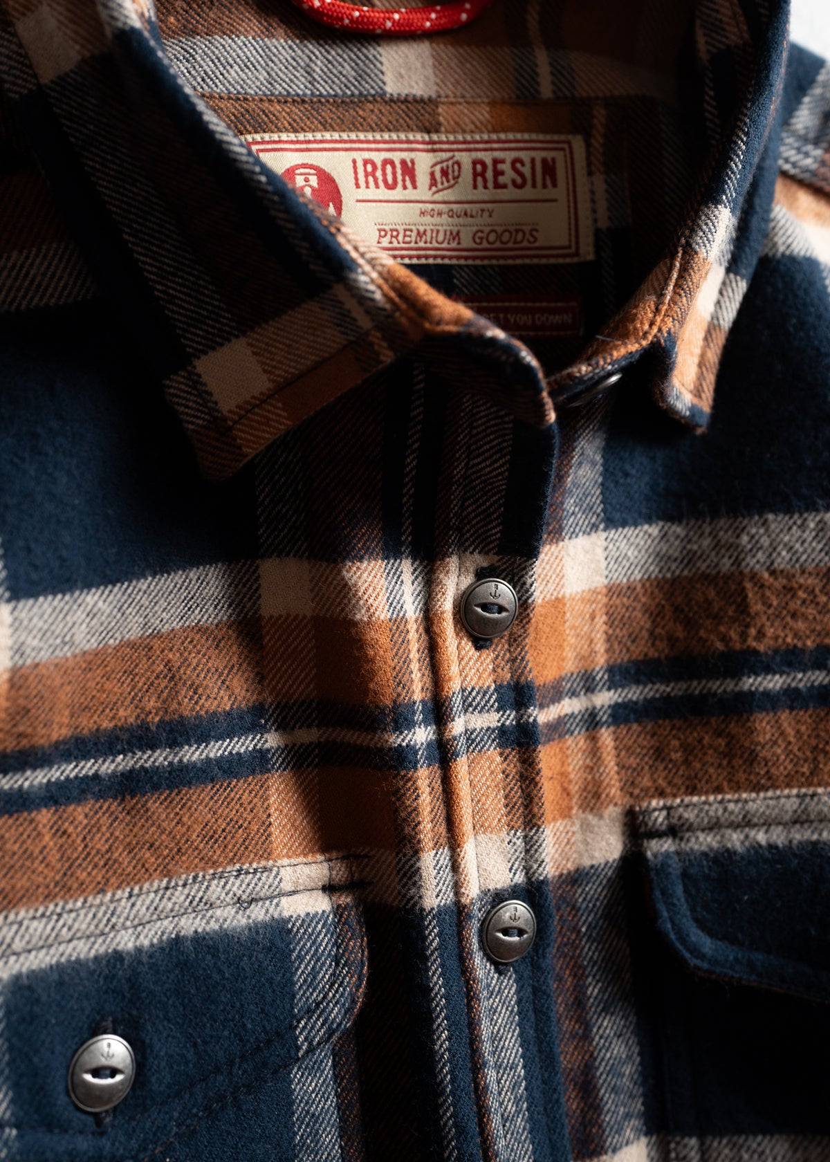 Close-up of a plaid shirt with brown, blue, and white checks. Visible label reads "Iron and Resin Premium Goods."